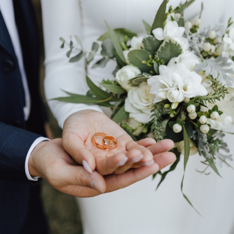 Wedding bands in the hands of bride and groom and with beautiful wedding bouquet made of greenery and white flowers